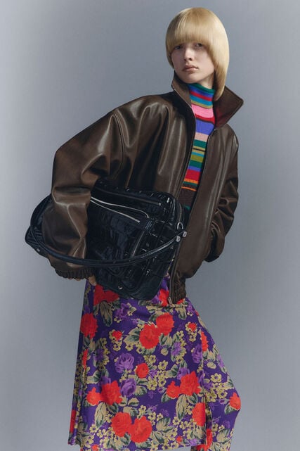 A fashion lookbook image of a female model wearing brown leather bomber jacket, floral printed skirt, knitted colourful turtle-neck sweater, holding a black leather Camero Camera shoulder bag.