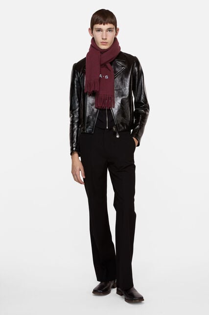 Male model wearing Acne Studios Canada fringe wool scarf in burgundy color, a small black leather biker jacket and black tailored pants.