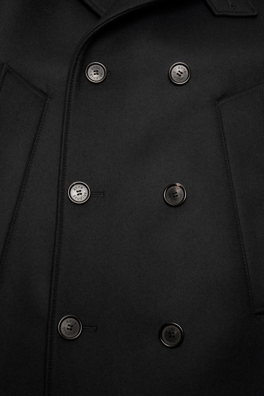 Close-up of an Acne Studios black wool peacoat, showing double-breasted front with six branded metal buttons and welt pocket detail.