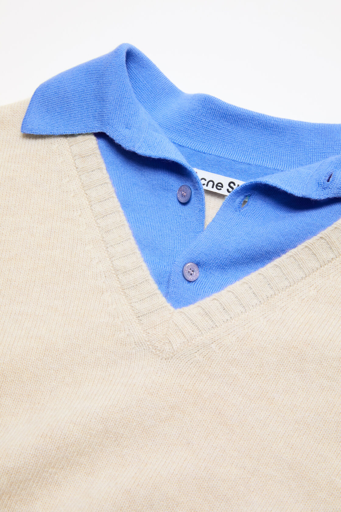 Close-up of an Acne Studios dirty white fine knit sweater with a V-neck, layered over a blue polo-collar underlayer with small buttons.