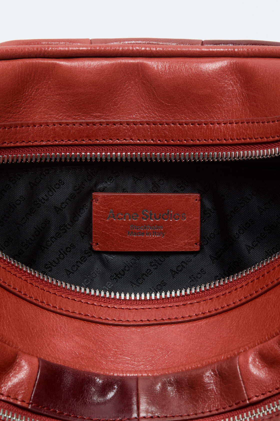Close-up of an Acne Studios burgundy leather double zipper closure with two silver-tone branded pull tabs and topstitched detailing.