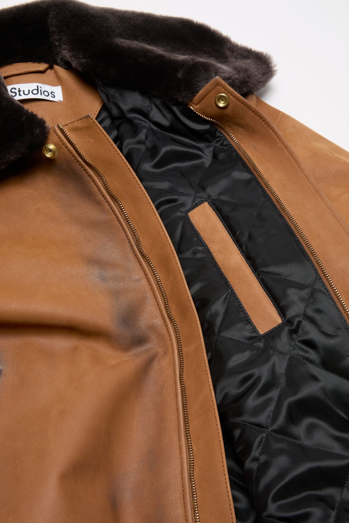 Close-up of an Acne Studios cognac brown suede bomber jacket, open to reveal a black quilted lining and an interior zip pocket, with a dark shearling