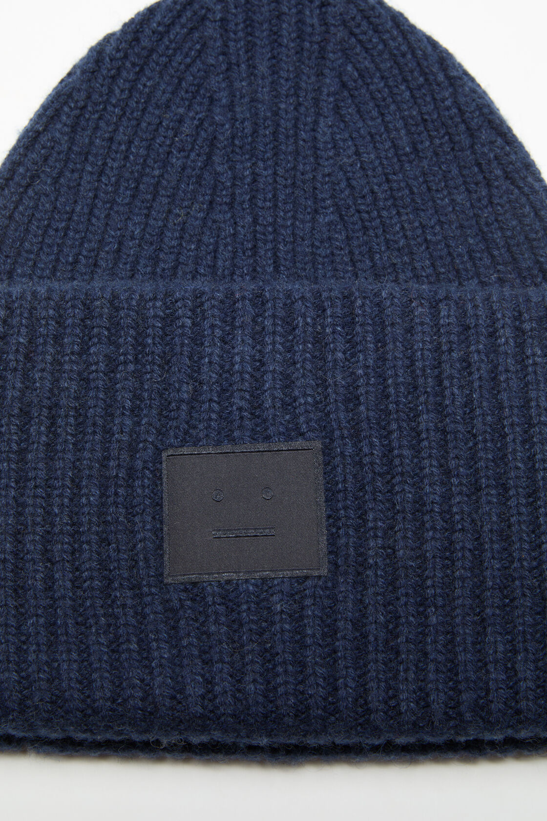 Close-up of an Acne Studios navy melange ribbed yak wool beanie hat with a folded cuff and a dark face logo patch.