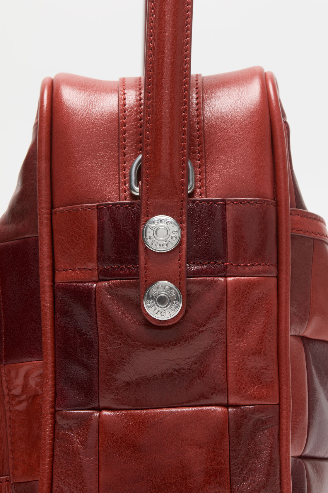 Close-up of an Acne Studios Camero Camera Patchwork Bag Bag in burgundy leather with silver zip compartments, button logo details, and a branded key