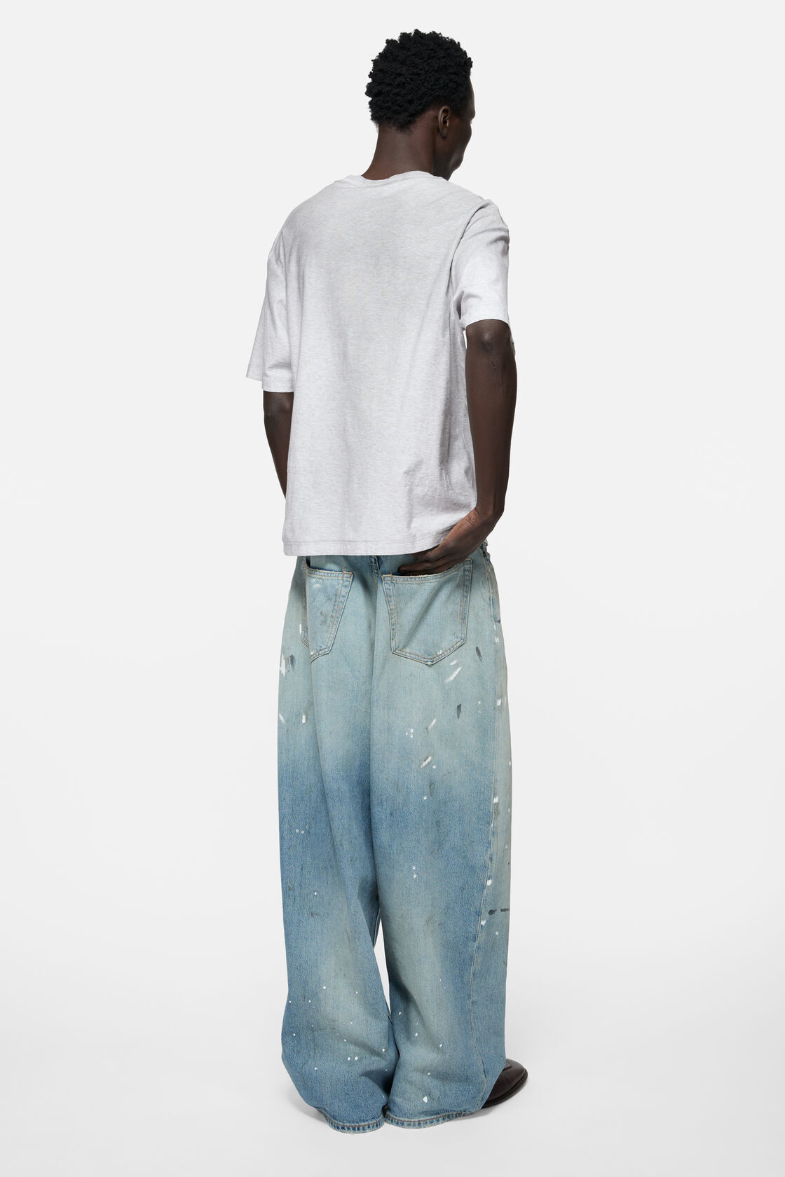Acne Studios pale grey melange sprayed logo t-shirt, relaxed fit, worn layered over a checked shirt with black wide-leg trousers and a blue denim bag.