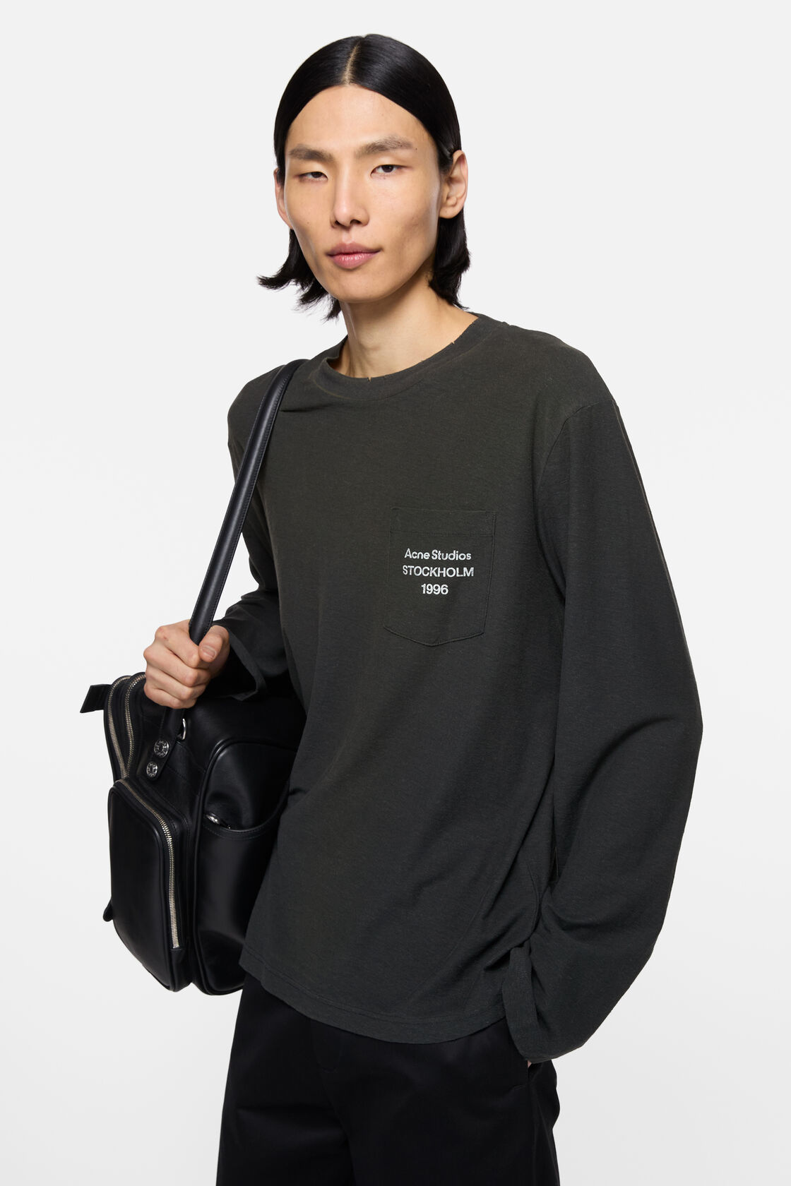 Acne Studios 褪色黑色长袖logo T恤，配有胸前口袋、圆领及宽松版型，搭配浅灰色长裤穿着。