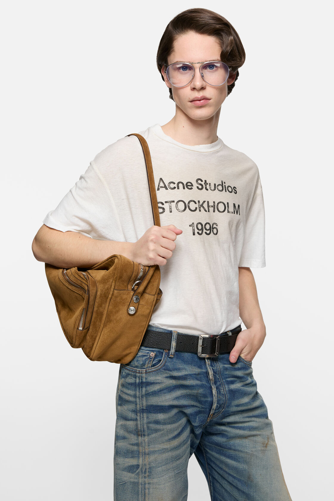 Acne Studios dusty white sprayed logo t-shirt with crewneck, worn with blue denim jeans, a black belt, and a tan suede shoulder bag.