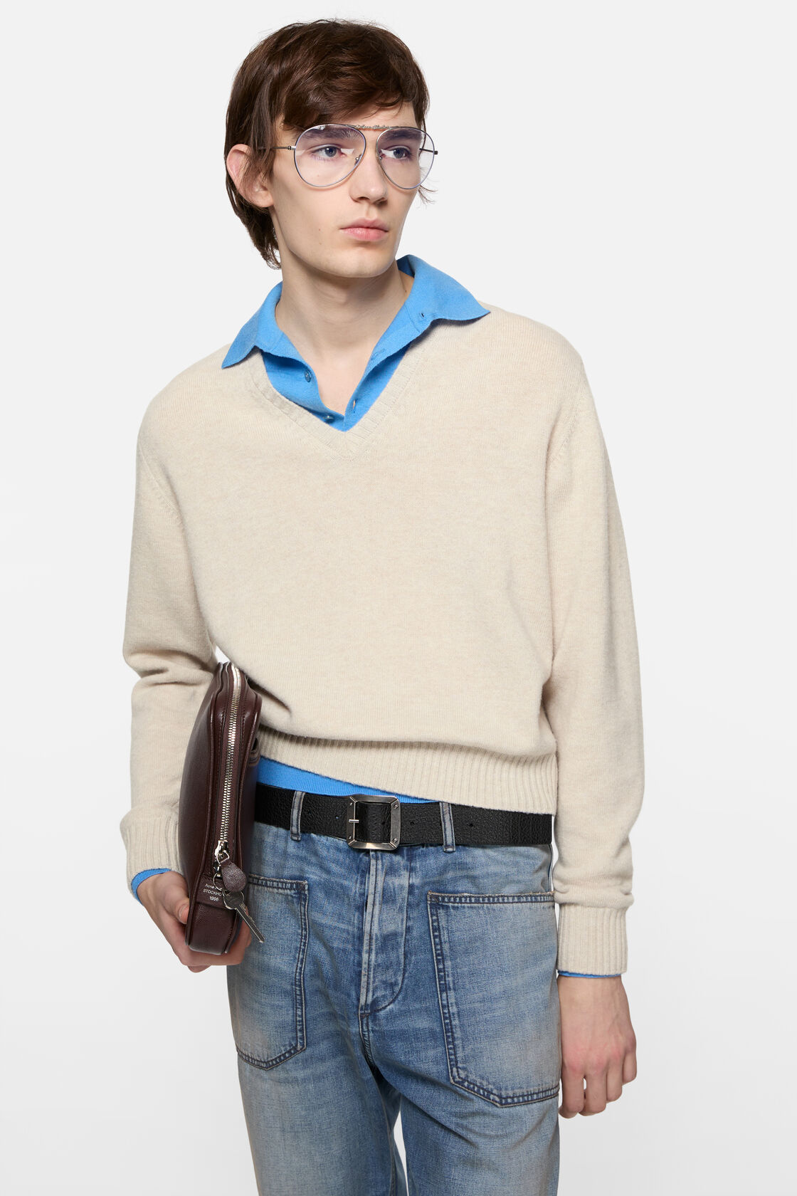 Acne Studios dirty white wool V-neck knit sweater layered over a blue polo shirt, styled with light-wash jeans, a black belt, and a dark brown zip