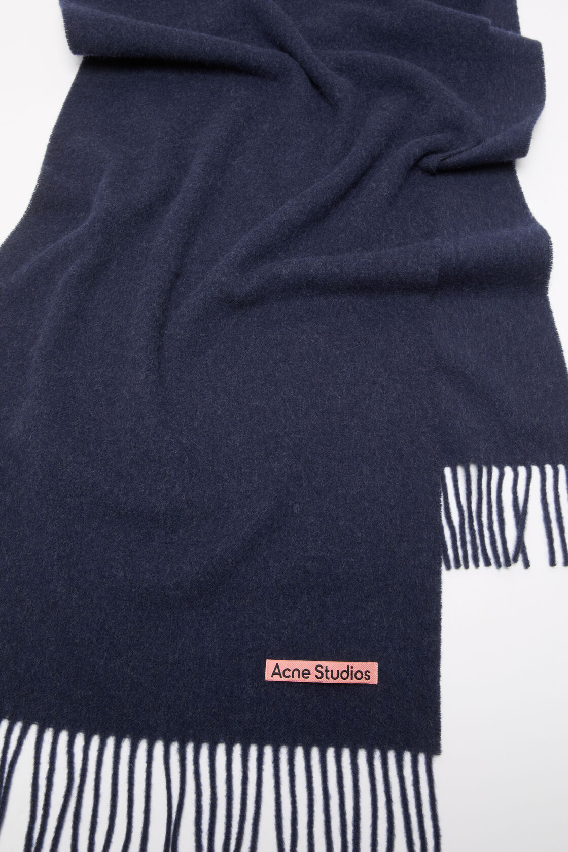 Acne Studios narrow fringe wool scarf in dark navy melange, laid flat showing fringed ends and a pink woven logo label.