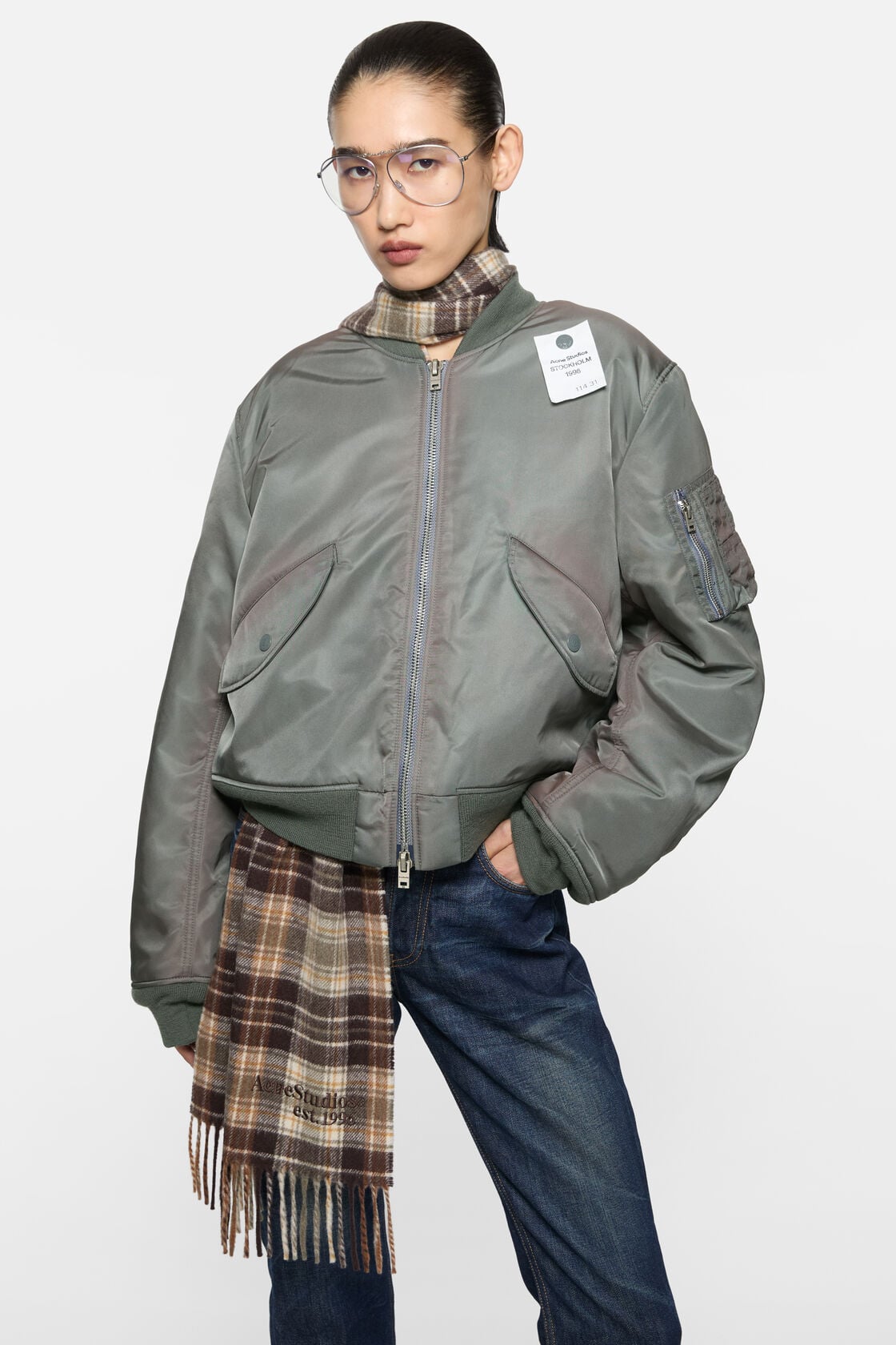 Acne Studios stone grey washed bomber jacket in recycled nylon, worn over a brown plaid fringed scarf and dark blue jeans.