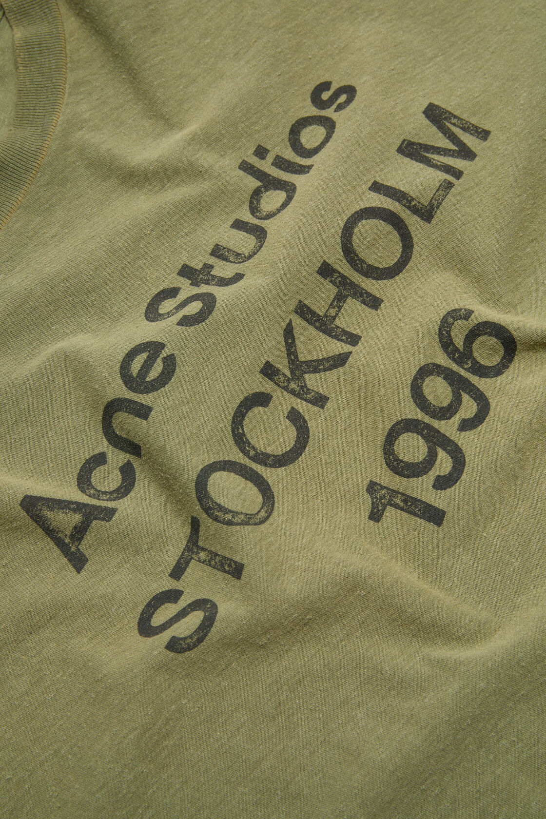 Close-up of an Acne Studios olive green sprayed logo t-shirt featuring a distressed 'Acne Studios Stockholm 1996' graphic print.