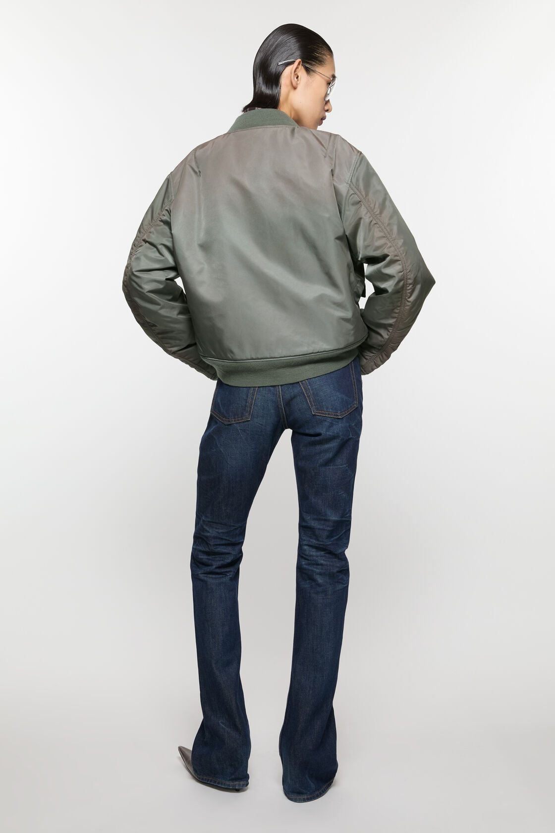 Acne Studios stone grey washed bomber jacket in recycled nylon, back view, worn with dark blue flare jeans and brown boots.