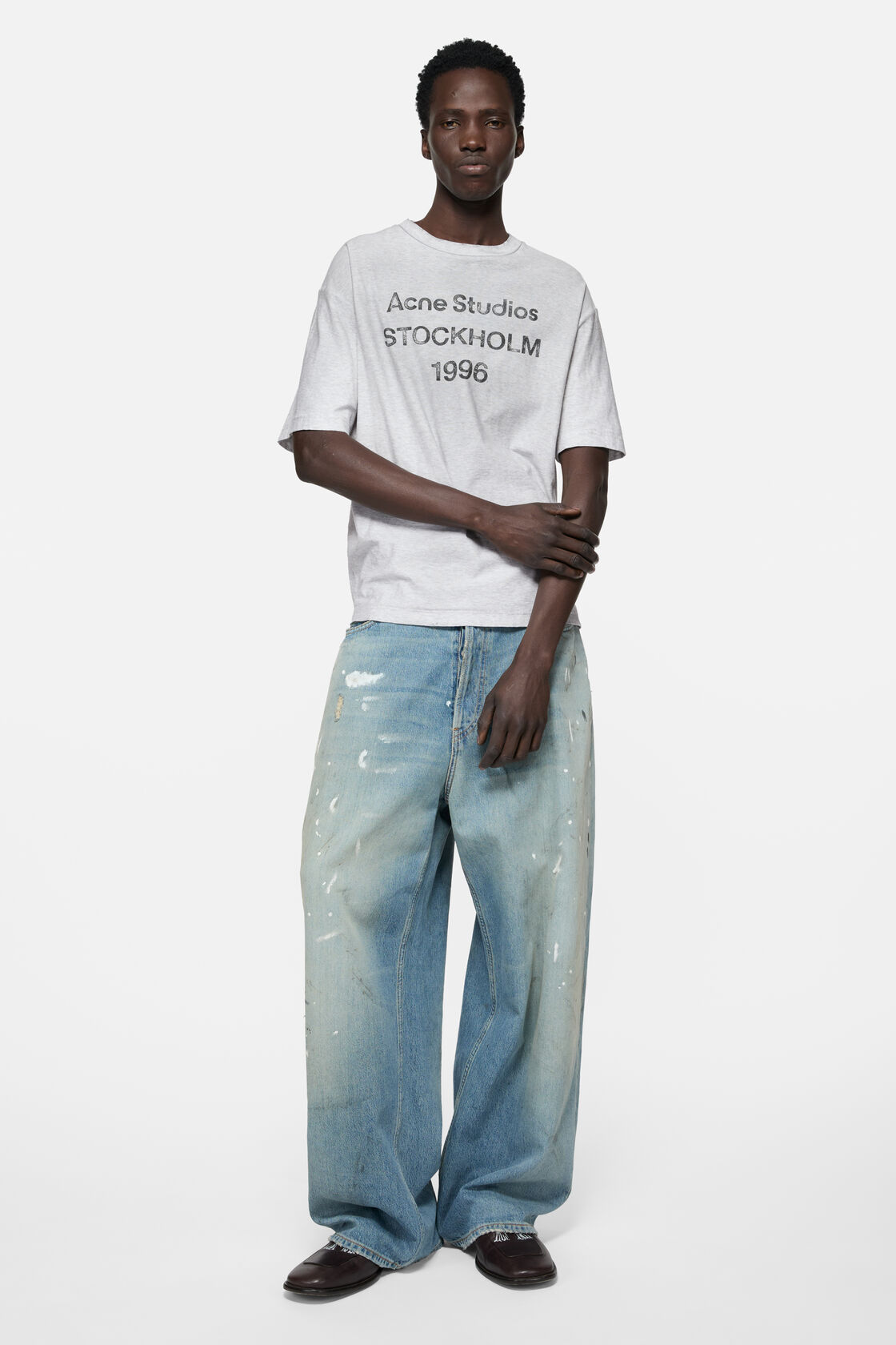Acne Studios pale grey melange sprayed logo t-shirt in a relaxed fit, worn over a checked long-sleeve shirt with black wide-leg trousers.