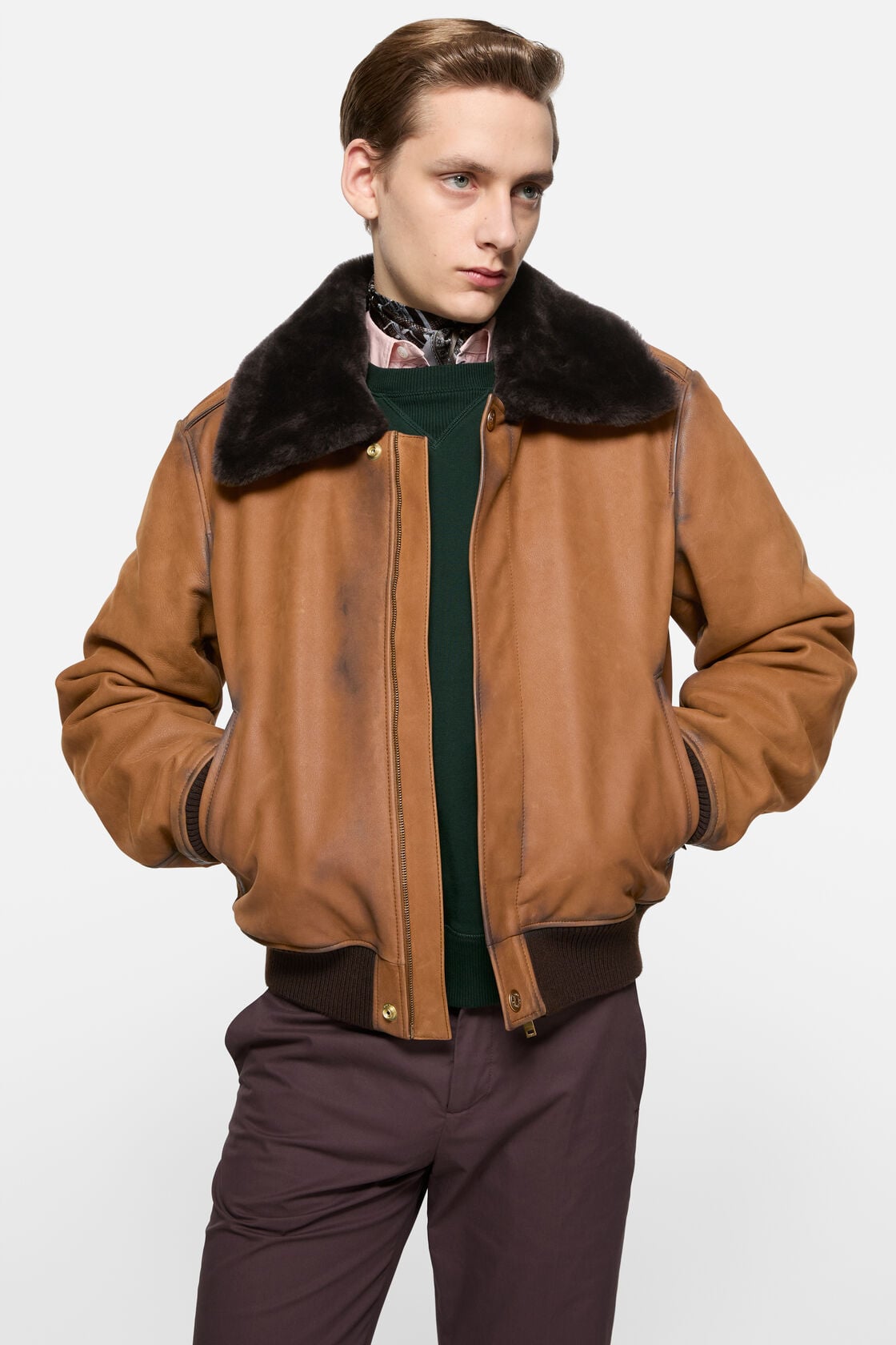 Acne Studios cognac brown suede bomber jacket with dark shearling collar, worn open over a green sweater and dark trousers.