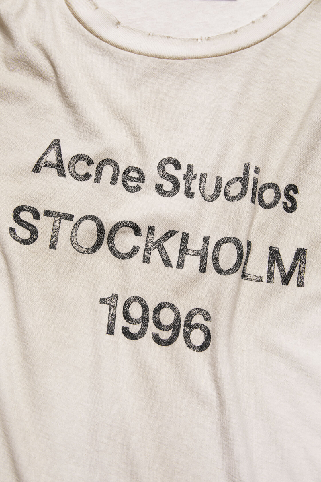 Close-up of an Acne Studios dusty white t-shirt chest area showing a distressed printed logo reading Acne Studios Stockholm 1996.