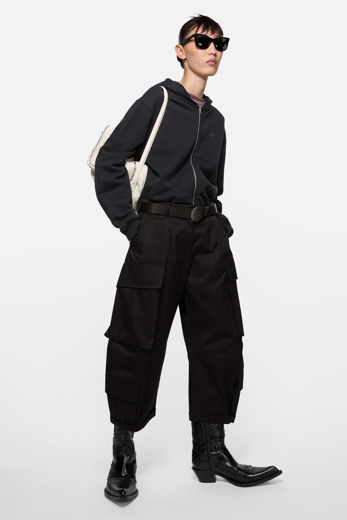 Acne Studios black leather belt worn with black cargo trousers, a dark zip-up hoodie, black cowboy boots, and a white tote bag.