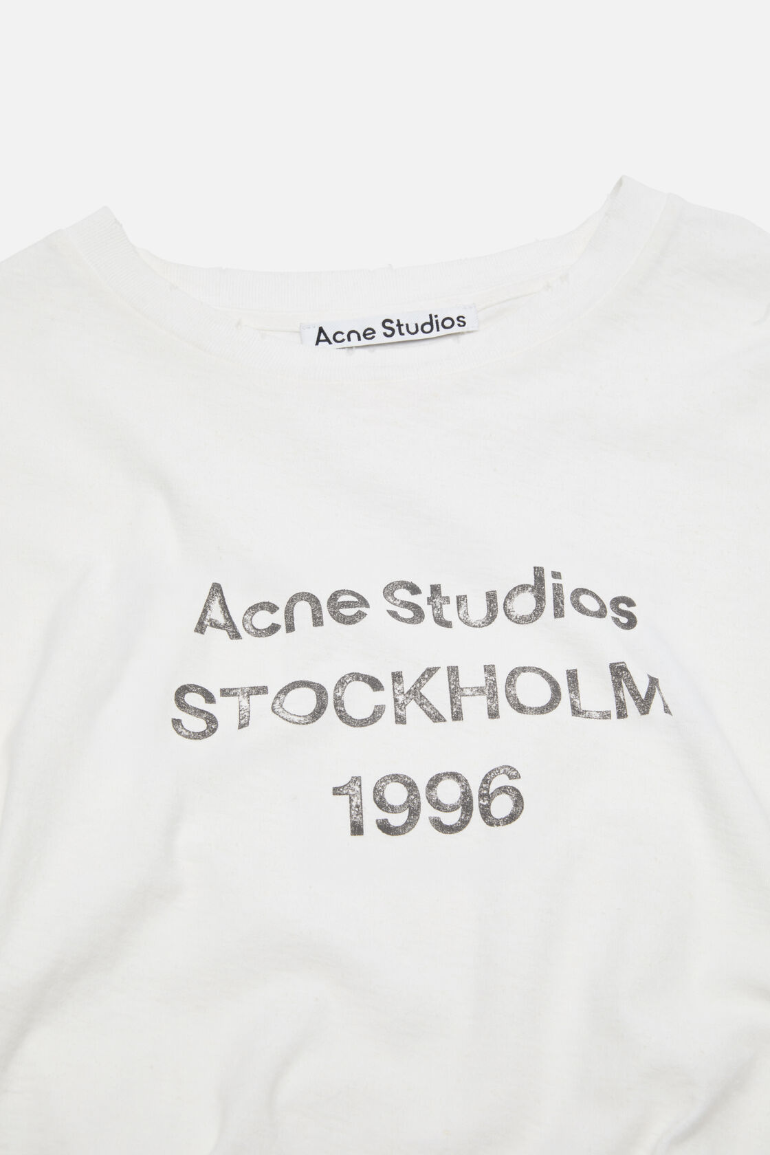 Close-up of an Acne Studios off-white logo t-shirt with silver distressed 'Acne Studios Stockholm 1996' graphic print at the front.