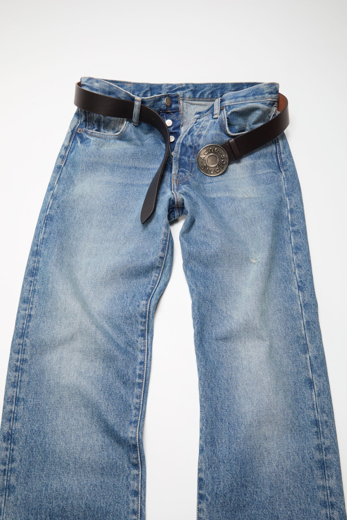Acne Studios black leather belt with a large circular antique silver logo buckle, styled through the loops of blue washed denim jeans.