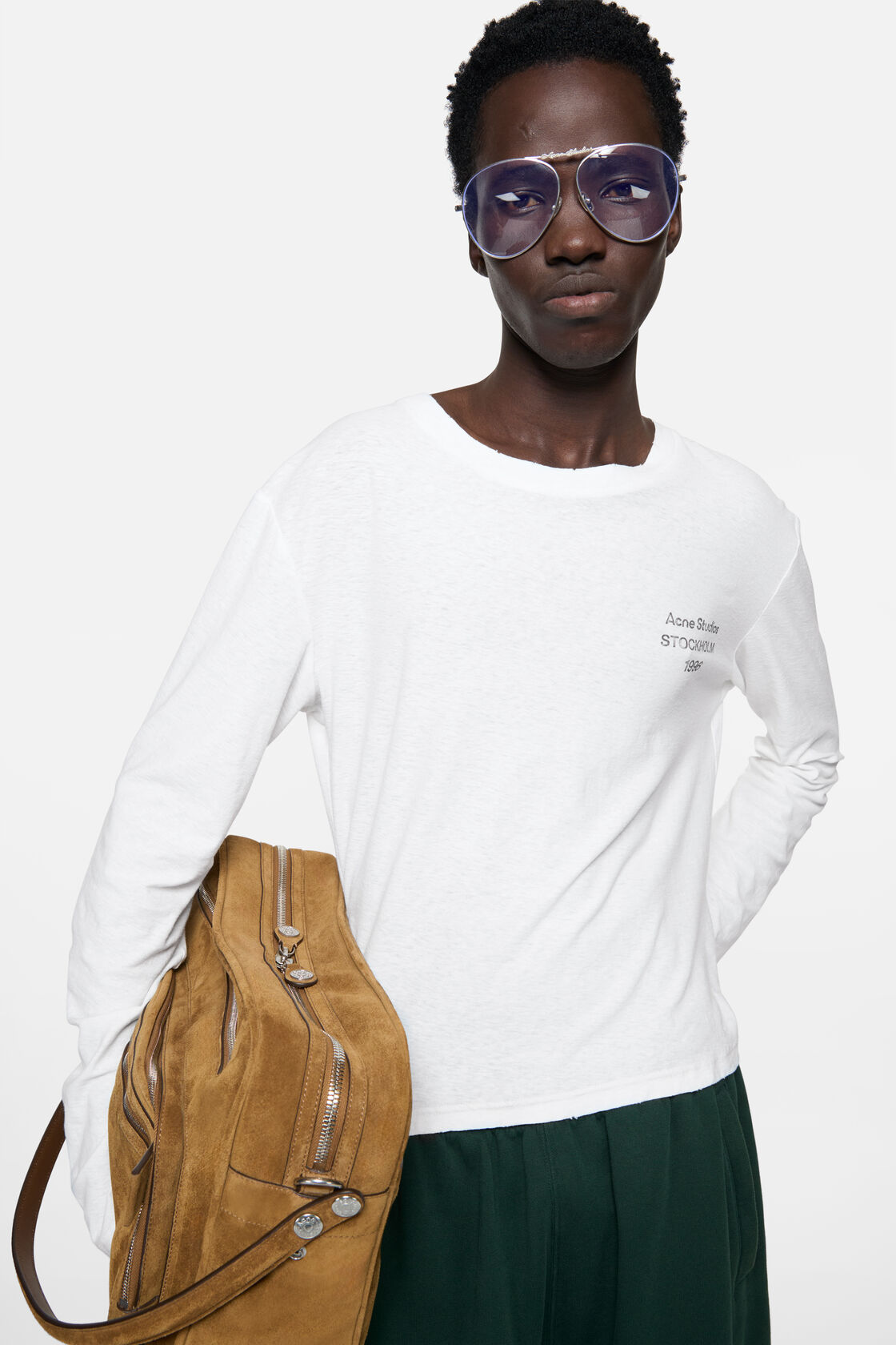 Acne Studios off-white distressed logo long-sleeve t-shirt in fitted silhouette, worn with dark green trousers, a tan suede bag, and tinted