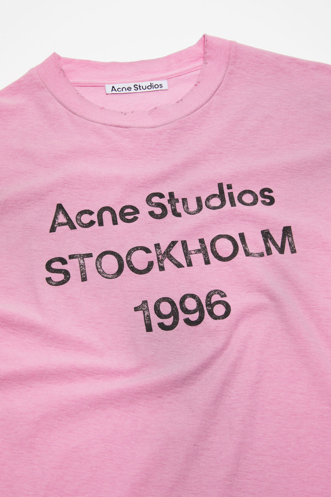 Close-up of an Acne Studios cotton candy pink crewneck t-shirt with a distressed spray-effect finish and printed Stockholm 1996 logo on the front.