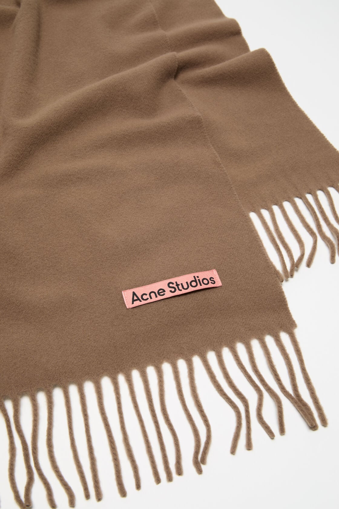 Close-up of an Acne Studios narrow fringe wool scarf in caramel brown, with fringe ends and a pink woven logo label visible.