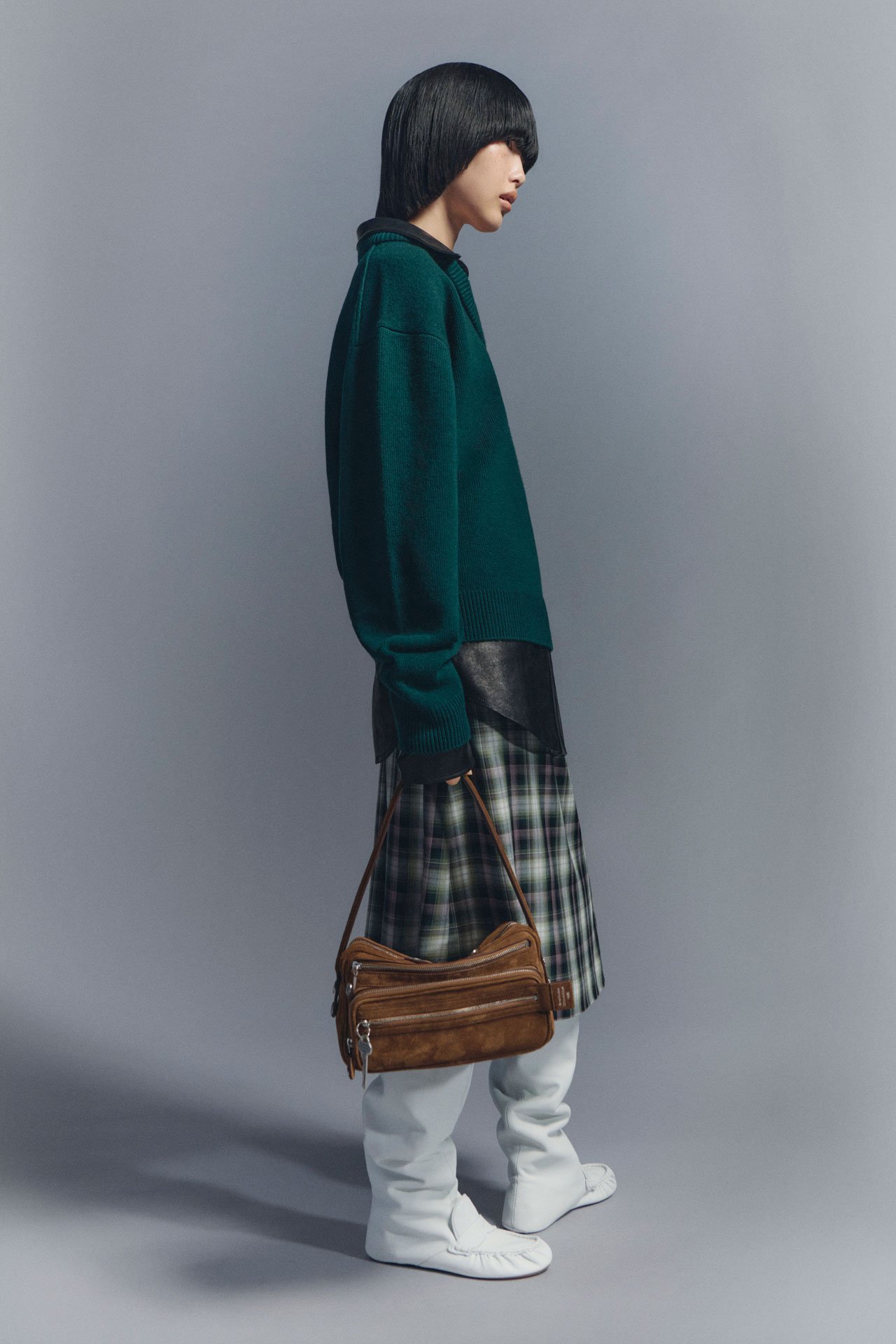 A fashion lookbook image of a female model wearing a forest-green knitted sweater, checked skirt, black blouse, white boots and holding a brown suede Camero bag.