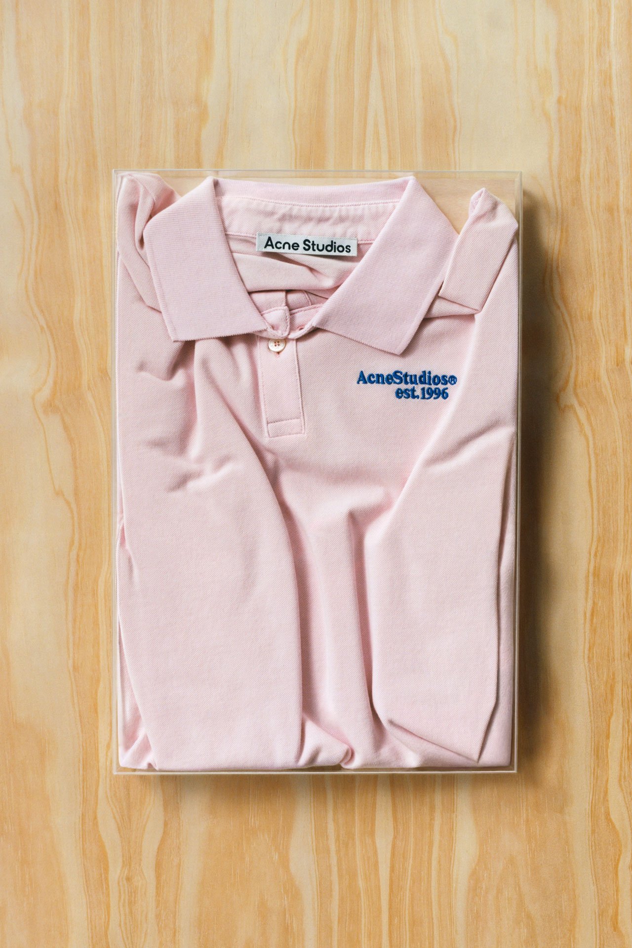 Acne Studios powder pink t-shirt with a blue logo on it folded on a wooden background.
