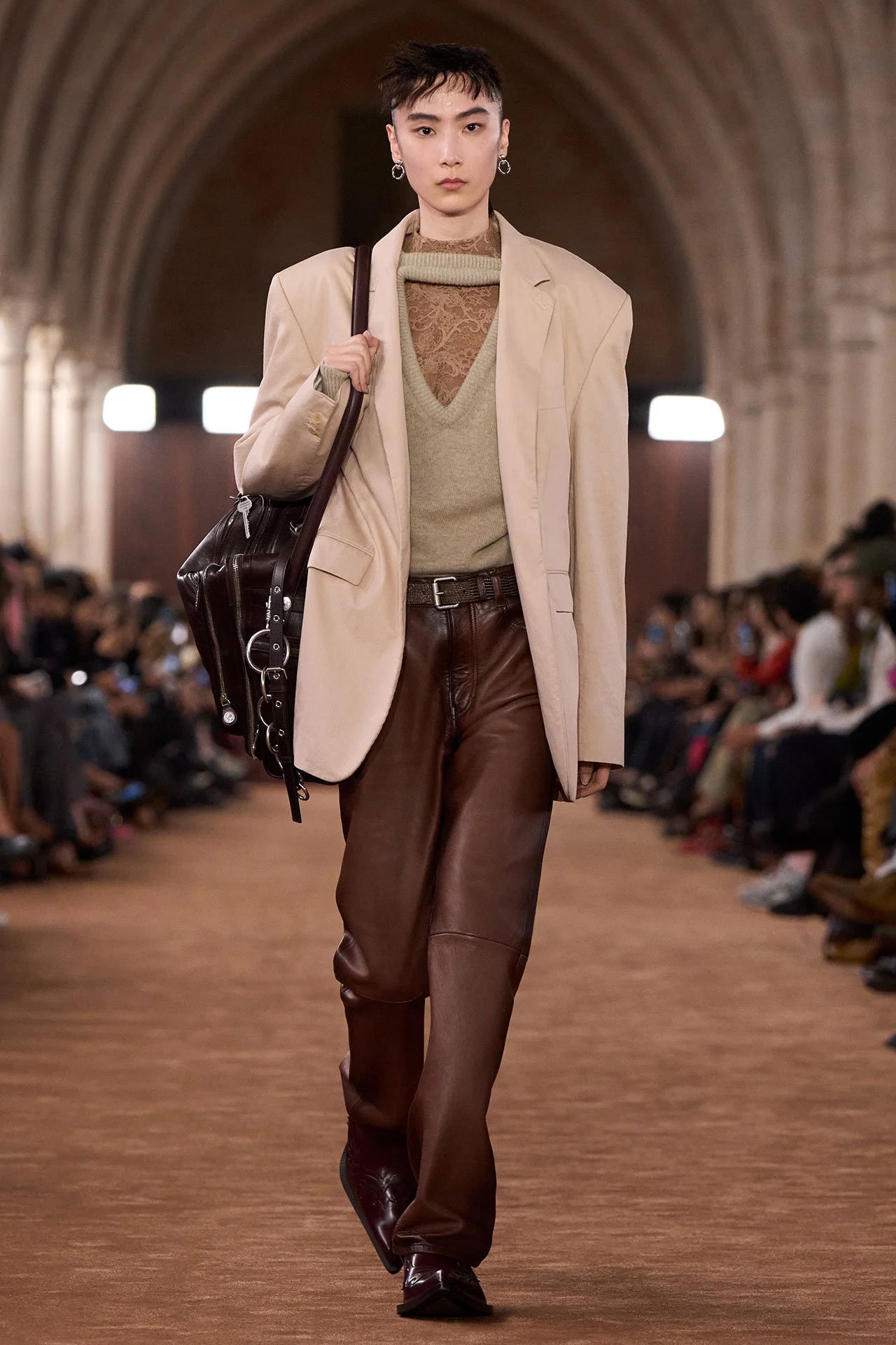 Acne Studios SS26 Runway - a female model wearing a beige suit jacket, beige knitted sweater, brown leather pants and holding a brown leather Camero bag.
