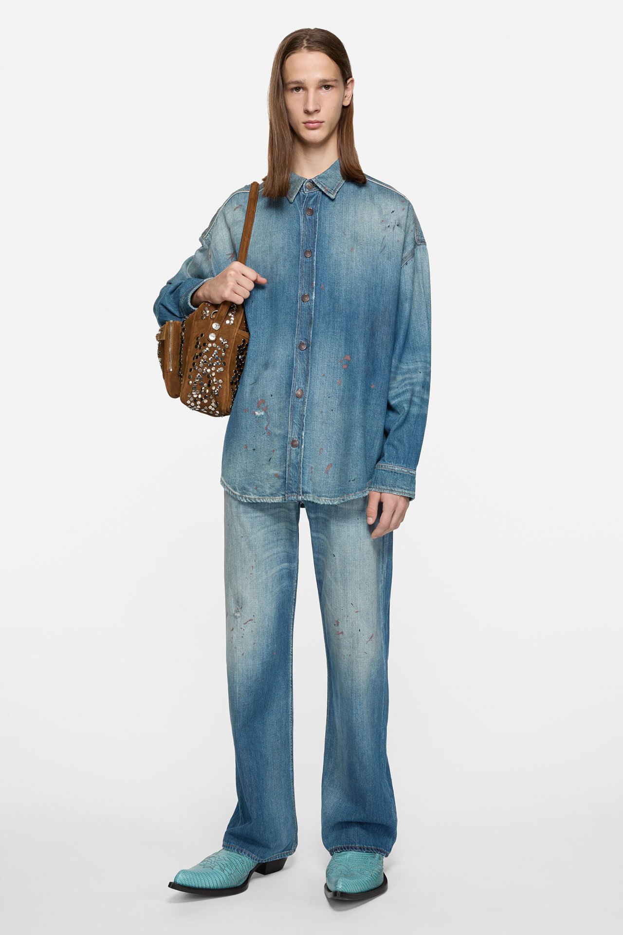 A male model wearing matching denim jeans and shirt, blue leather cowboy boots and brown suede Camero bag.