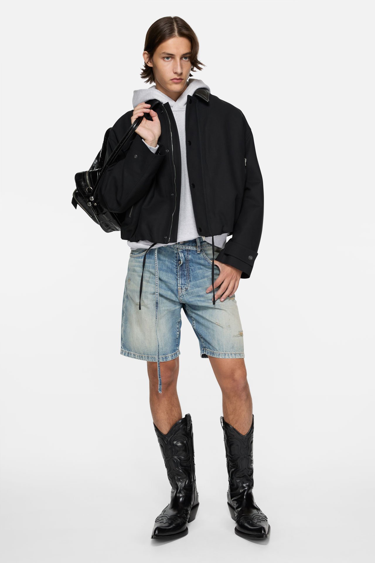 A male model wearing a short black bomber jacket grey hoodie, blue short jeans and black leather boots.