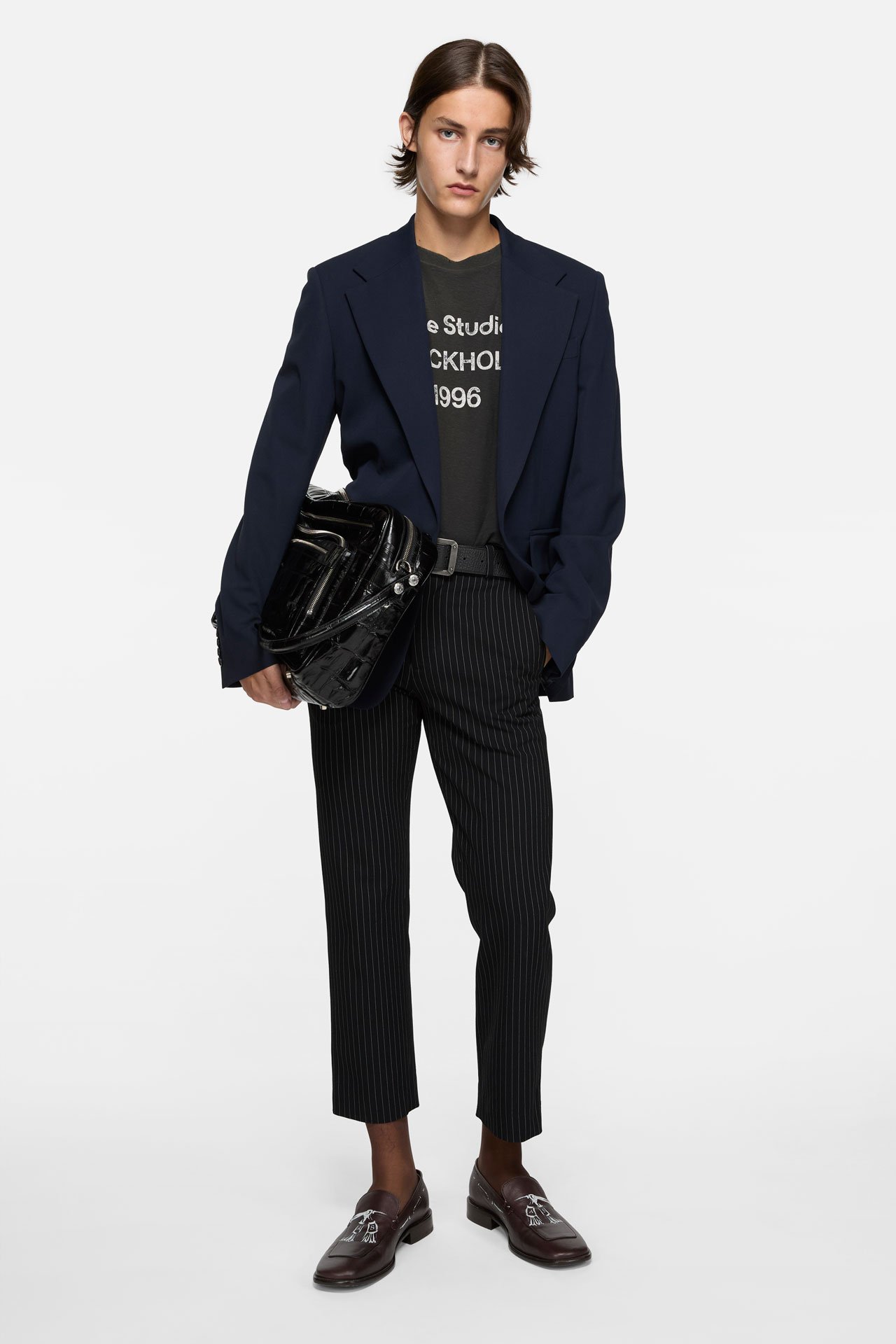 A male model wearing Acne Studios black ankle trousers, black 1996 logo t-shirt and a black tailored suit jacket, holding a black leather Camero bag.