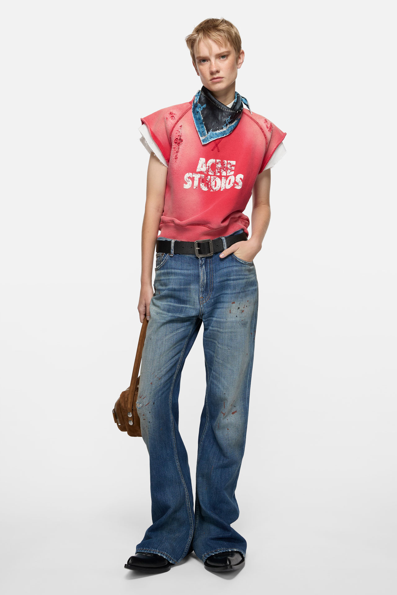 A female model wearing a pair of mid-blue Acne Studios jeans, short red t-shirt and black and blue scarf, holding a brown Camero bag.