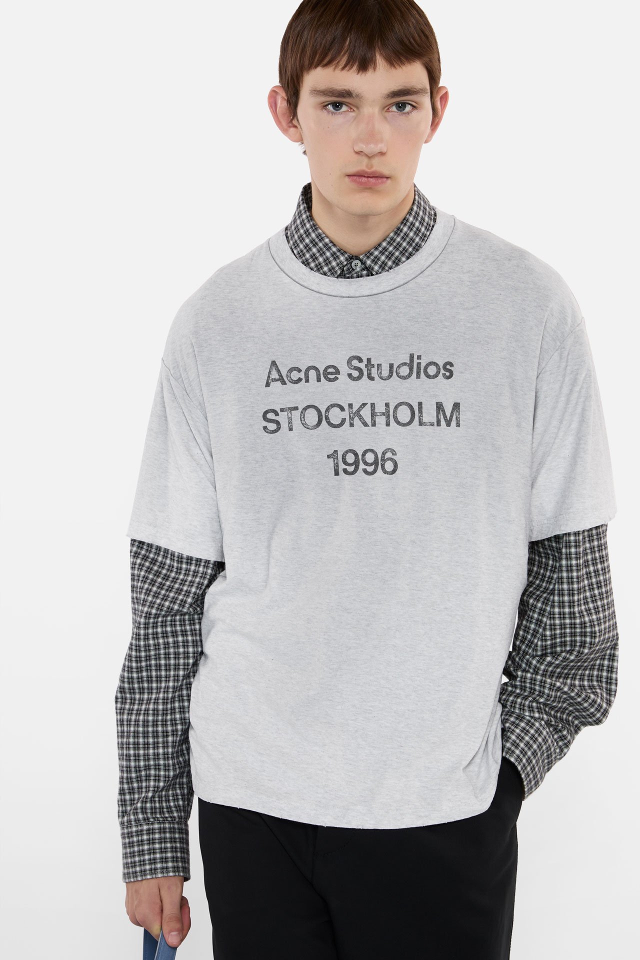 A male model wearing an Acne Studios 1996 logo light grey t-shirt on top of a grey checked long-sleeved shirt, wearing black pants.