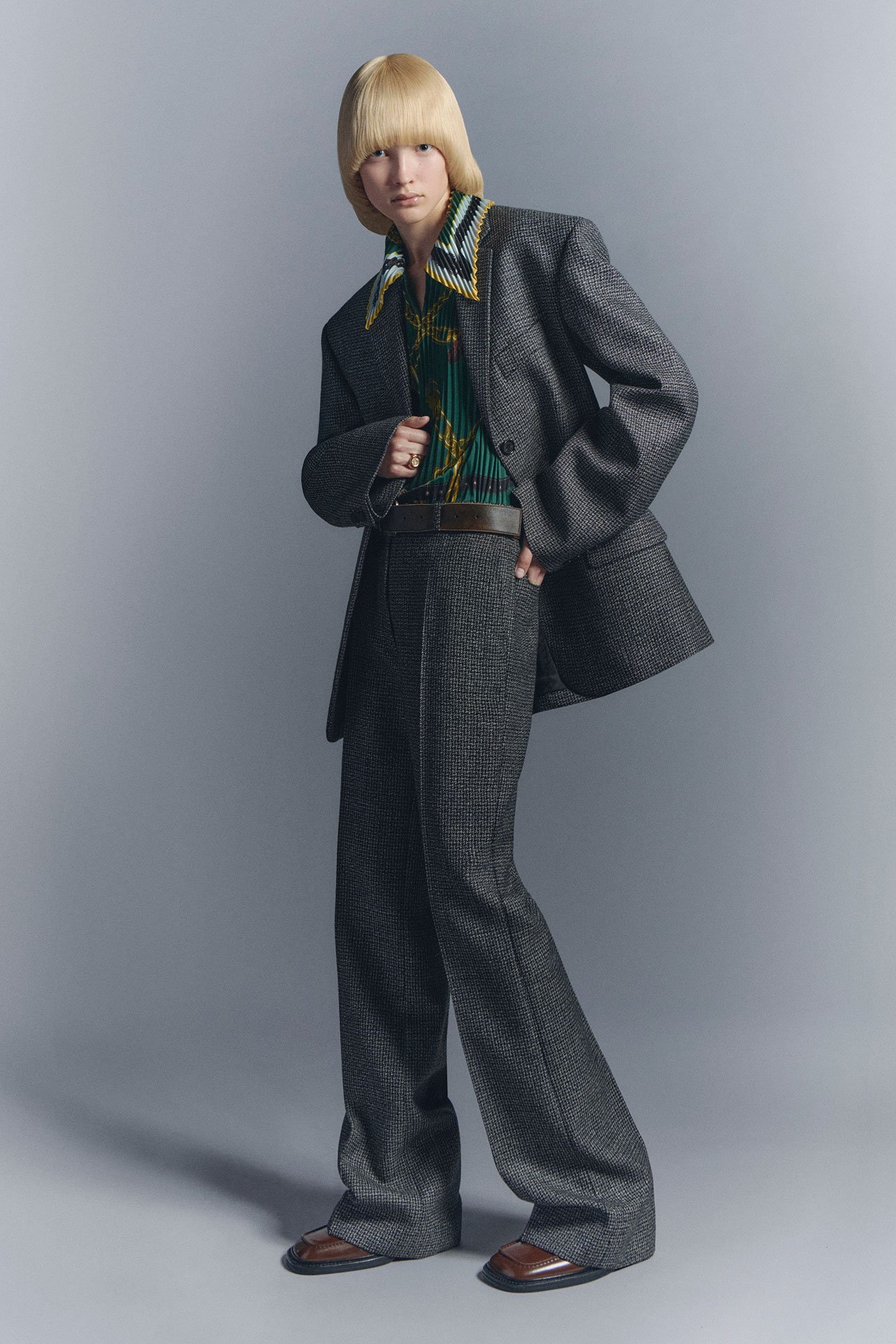 Blond woman in Acne Studios SS26 new arrivals grey tailored suit jacket and trousers, with brown leather shoes and belt, and a green shirt.