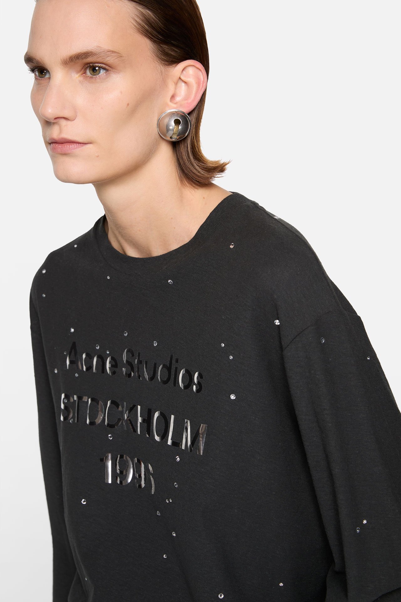 A close-up of female model showing Acne Studios black long-sleeve t-shirt with rhinestones, wearing a bold earring.