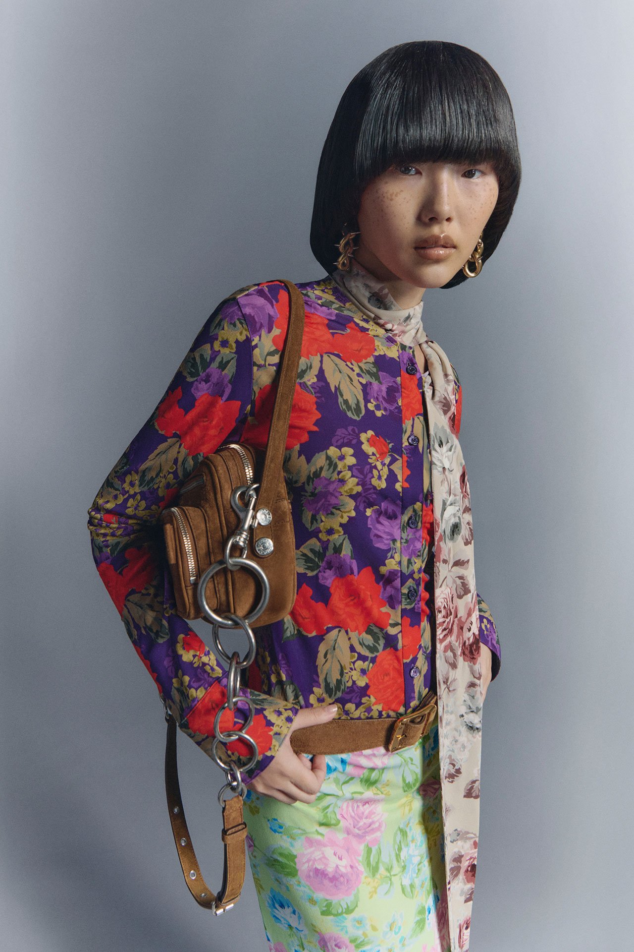 Female model wearing a a purple flower-print shirt and green flower-print skirt, having a brown suede Camero bag on her shoulder.