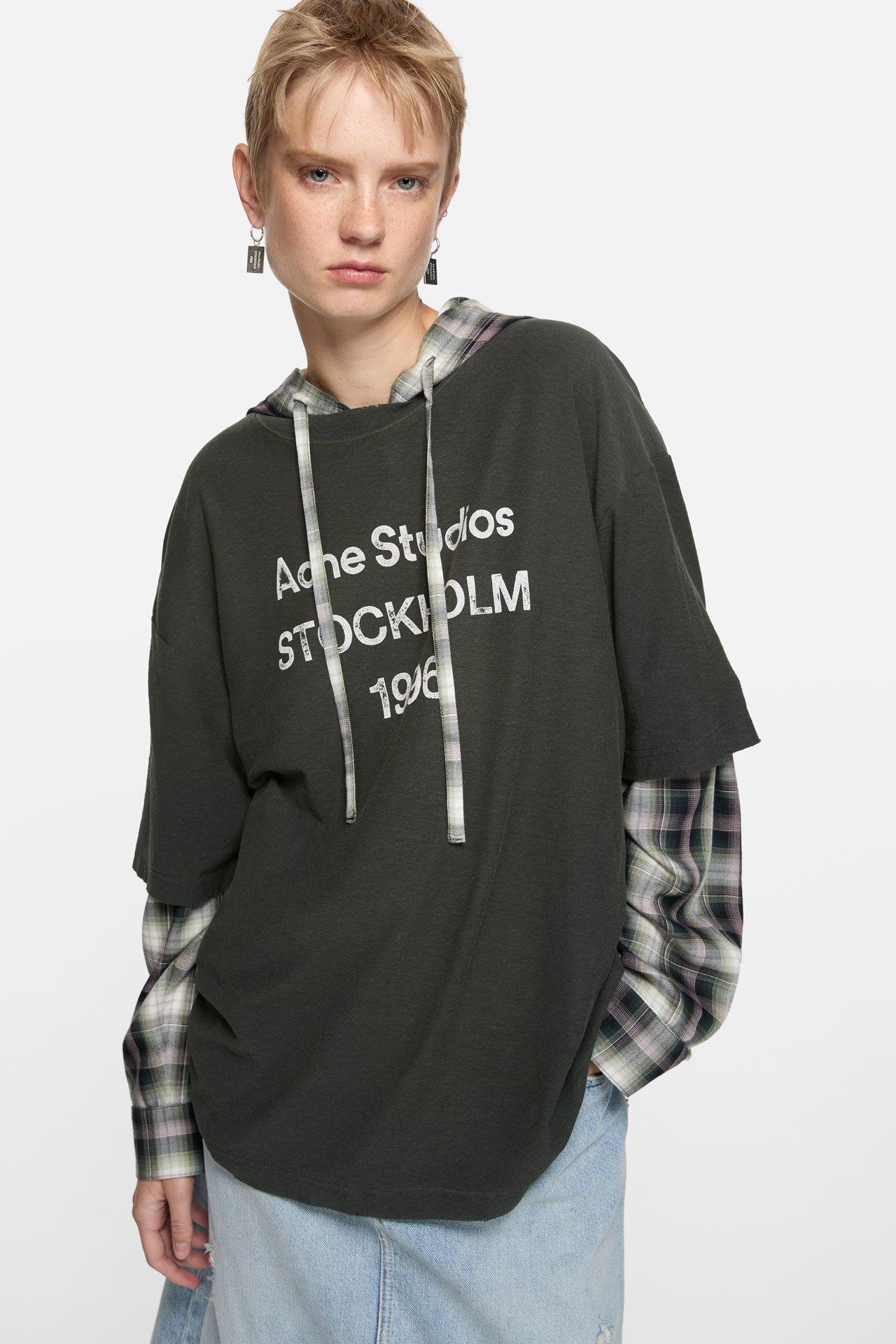A blond short-haired female model wearing an Acne Studios 1996 logo dark grey t-shirt on top of a grey checked long-sleeved shirt and blue jeans skirt.