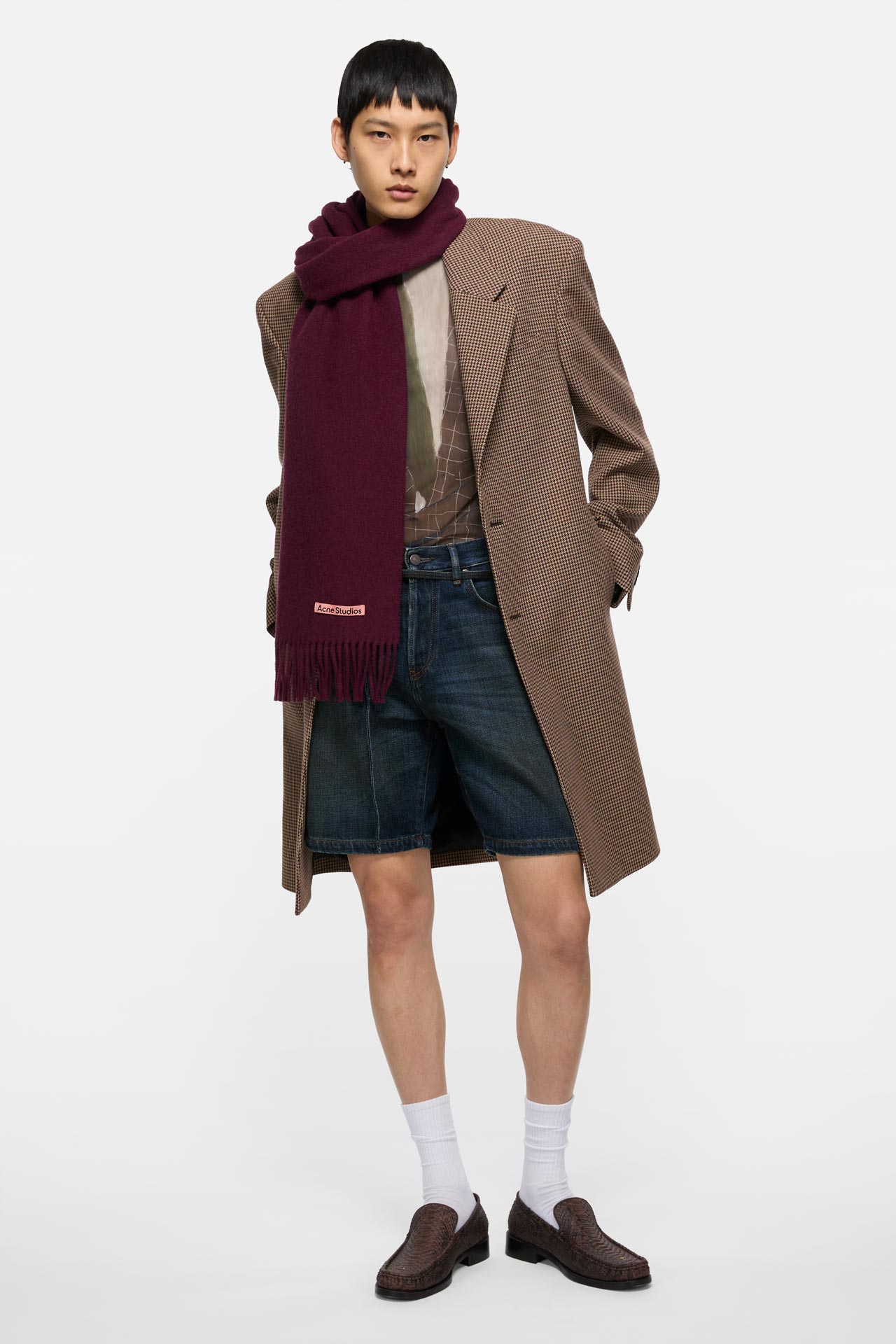 A male model wearing a Canada fringe wool scarf in Burgundy melange, long brown checked suit jacket, blue jeans shorts and brown crocodile loafers.