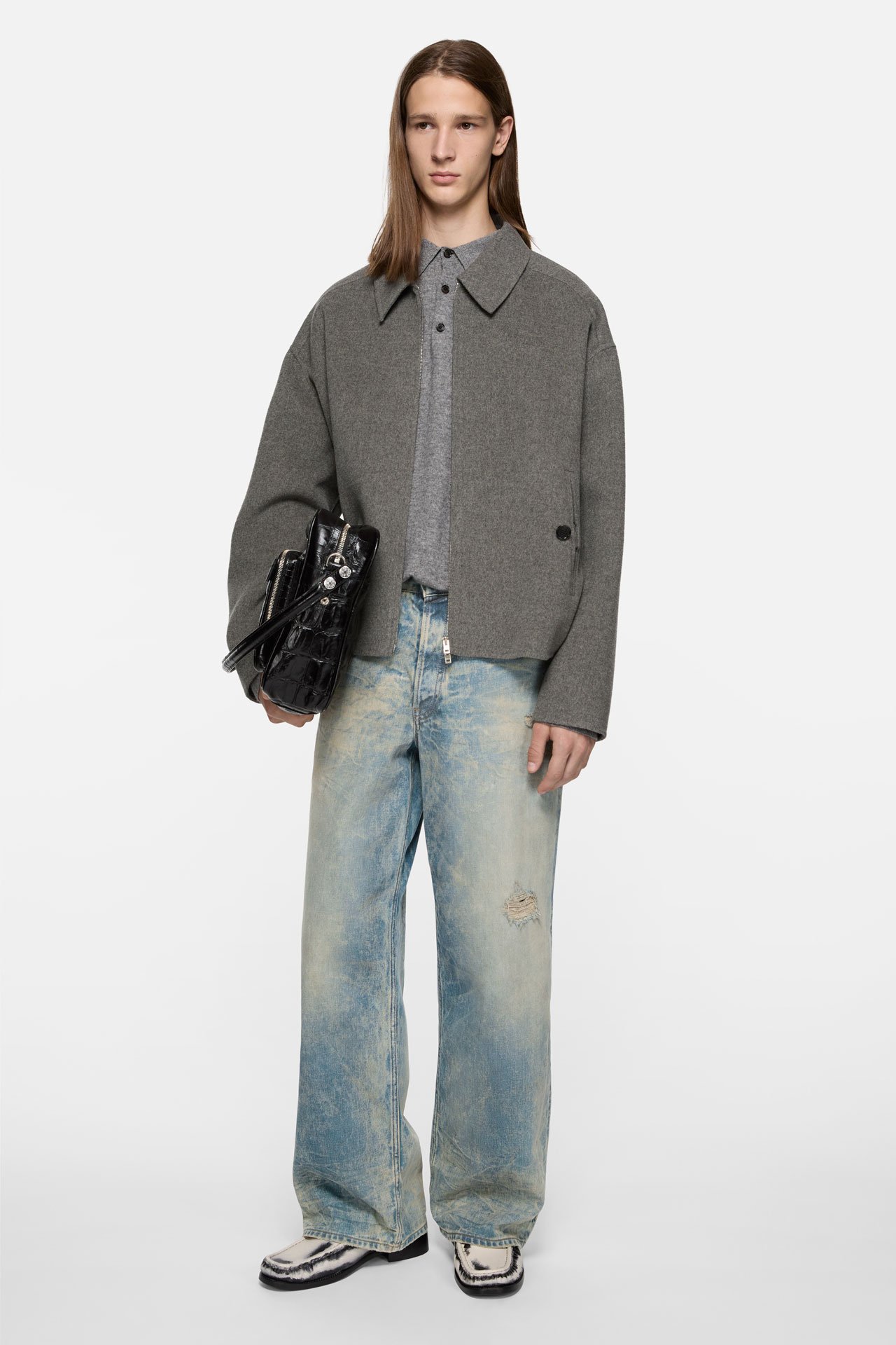 A male model wearing an Acne Studios light blue loose fit jeans in 1981 style, a grey cashmere jacket and a crockodile black leather Camero bag.