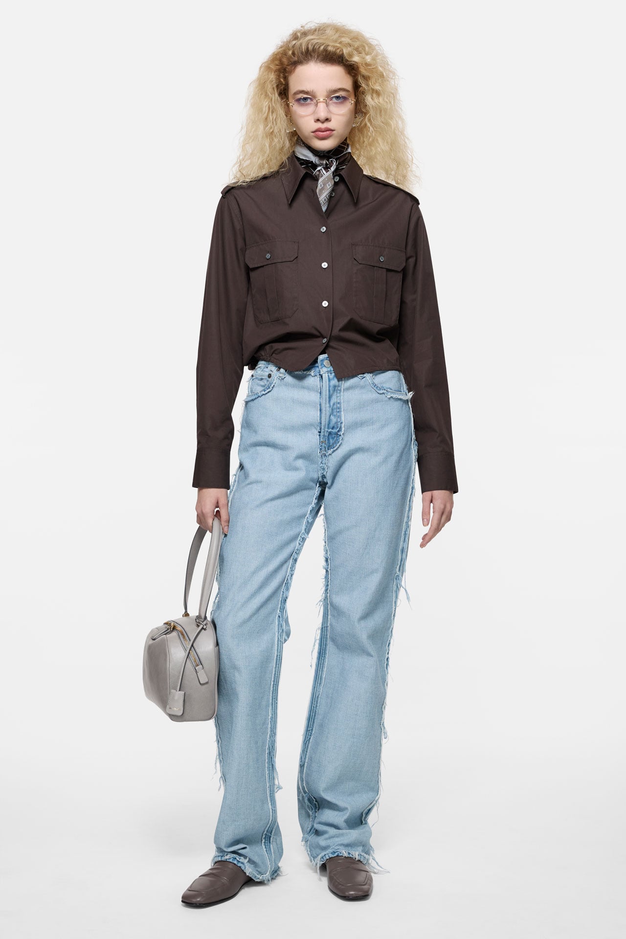 Acne Studios light blue jeans with brown bomber jacket, holding a grey leather bag.