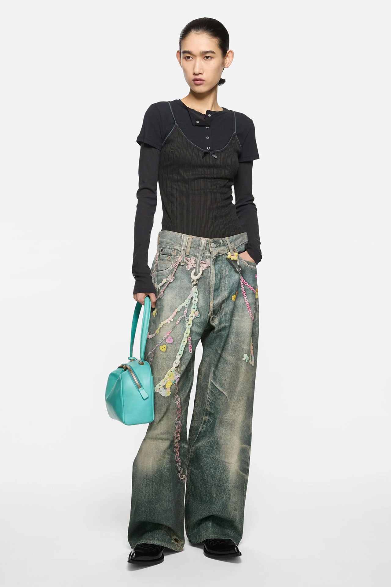 Woman wearing Acne Studios TLO keychain blue baggy jeans, black knitted long-sleeve layered top, and holding a bright blue leather bag.