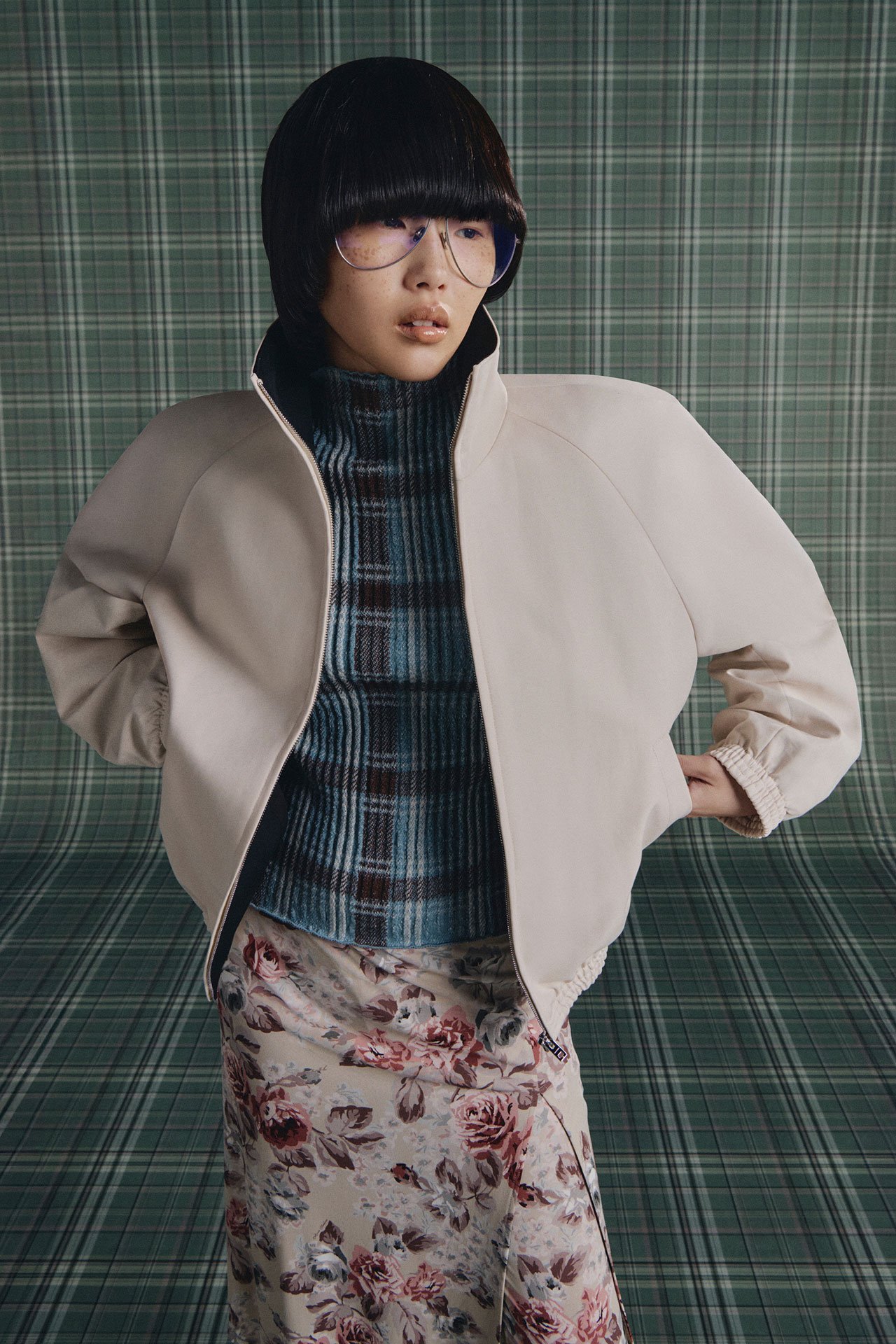 A close-up of a female model with glasses is wearing Acne Studios SS26 new arrivals beige bomber jacket, blue brown checked shirt and a skirt with roses in front of a green checked background..