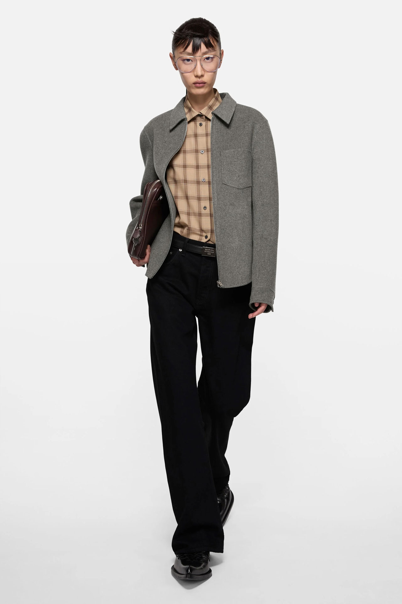 A female model wearing a grey cashmere zip jacket, beige checked shirt, black jeans and glasses.