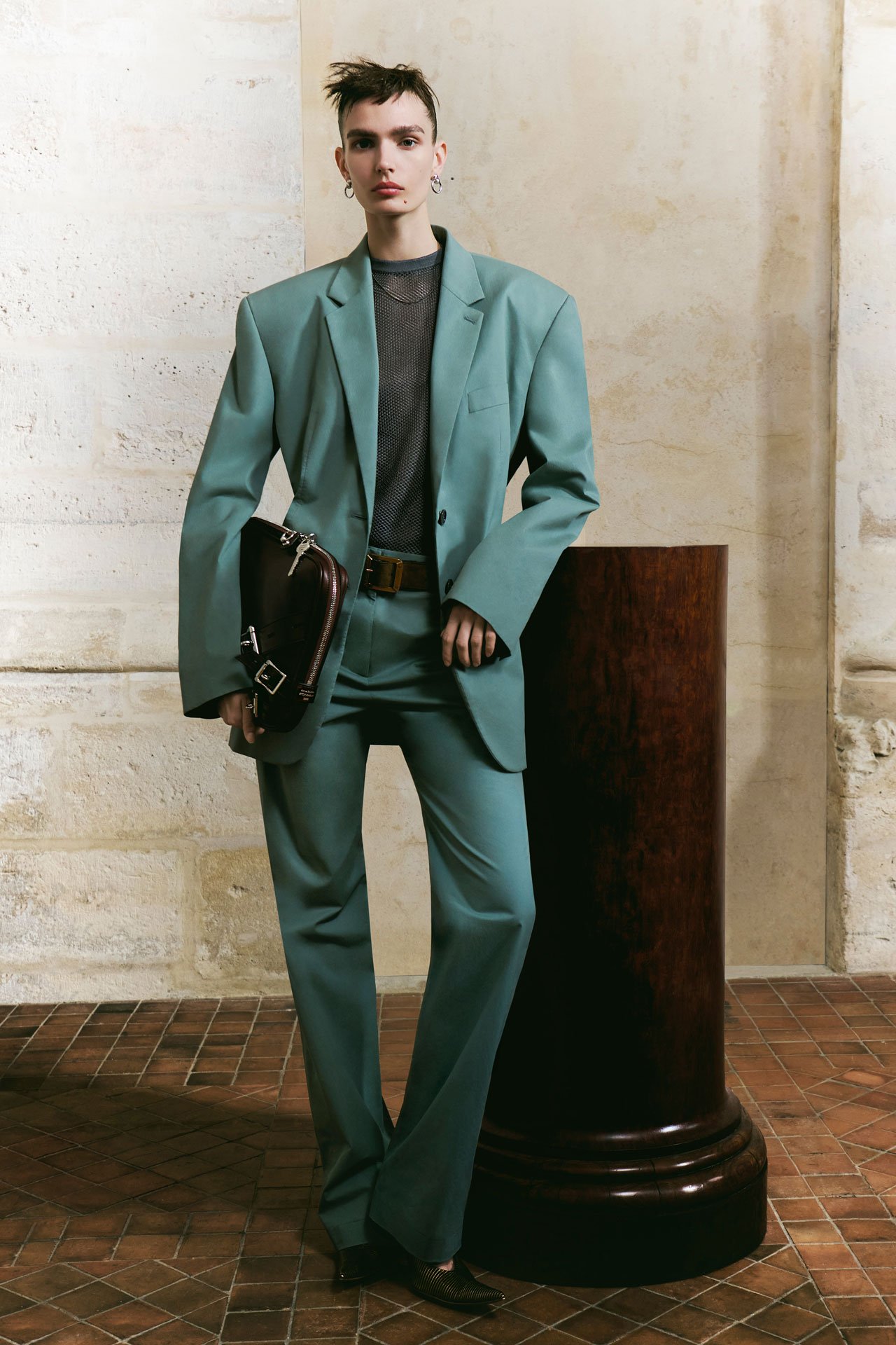 SS26 Acne Studios Paris Fashion Show female model wearing a blue matching tailored suite and pants, grey shirt and holding a brown leather laptop case.