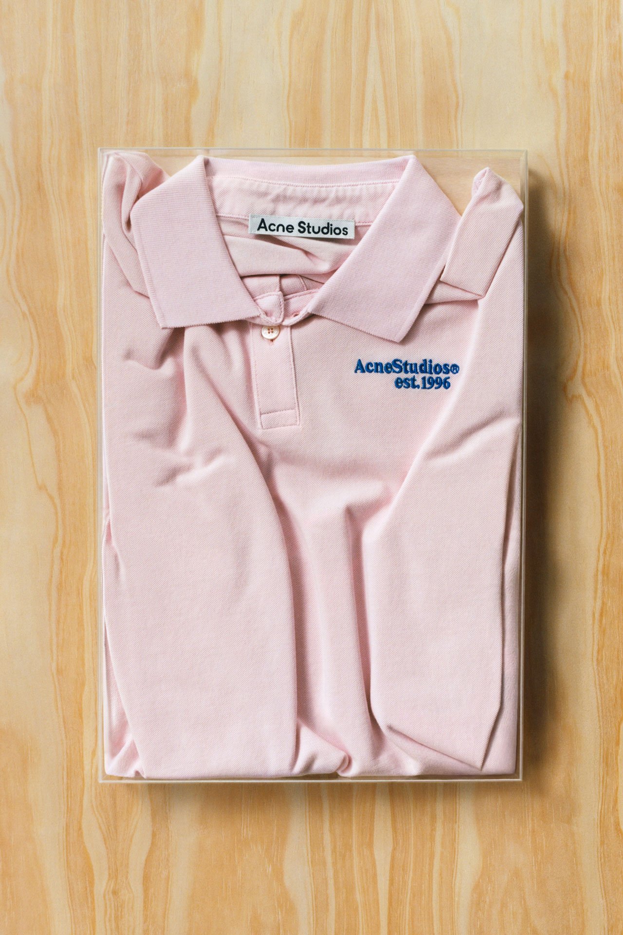 Acne Studios SS26 Summer Collection light pink t-shirt with collar and blue Acne Studios logo folded and presented on a light wooden tray.