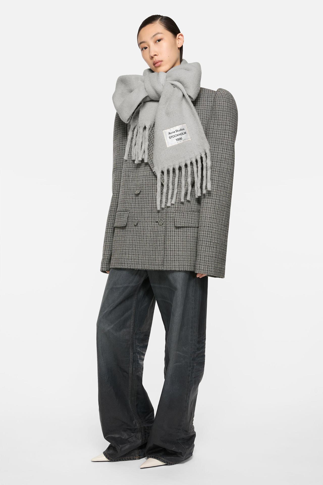Female model wearing an Acne Studios 1996 bicolour alpaca scarf in grey, padded-shoulder grey checked suit jacket and dark blue jeans.