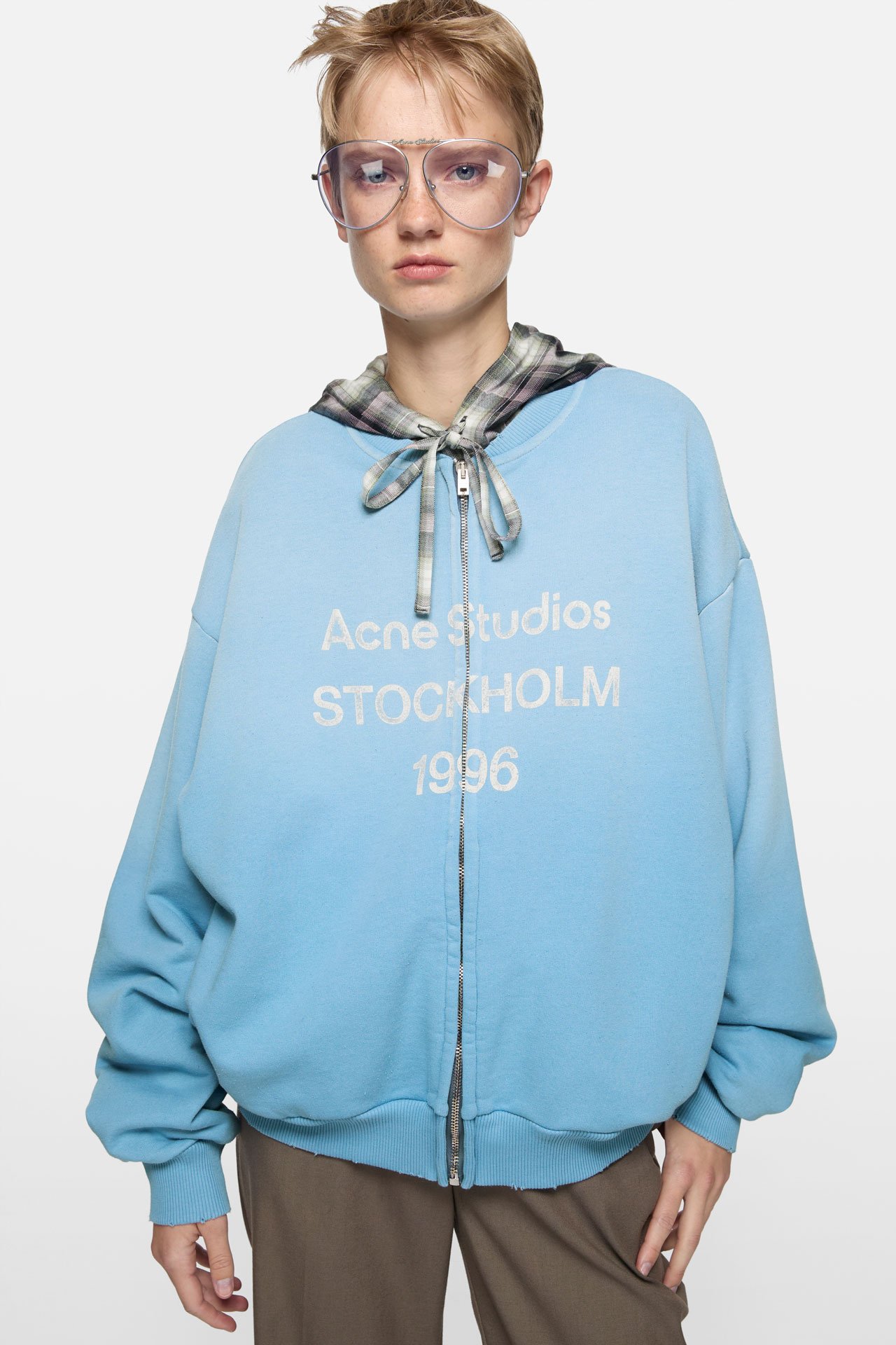 A female model wears a light blue sweater by Acne Studios with the signature 1996 logo on it, brown pants and clear glasses.
