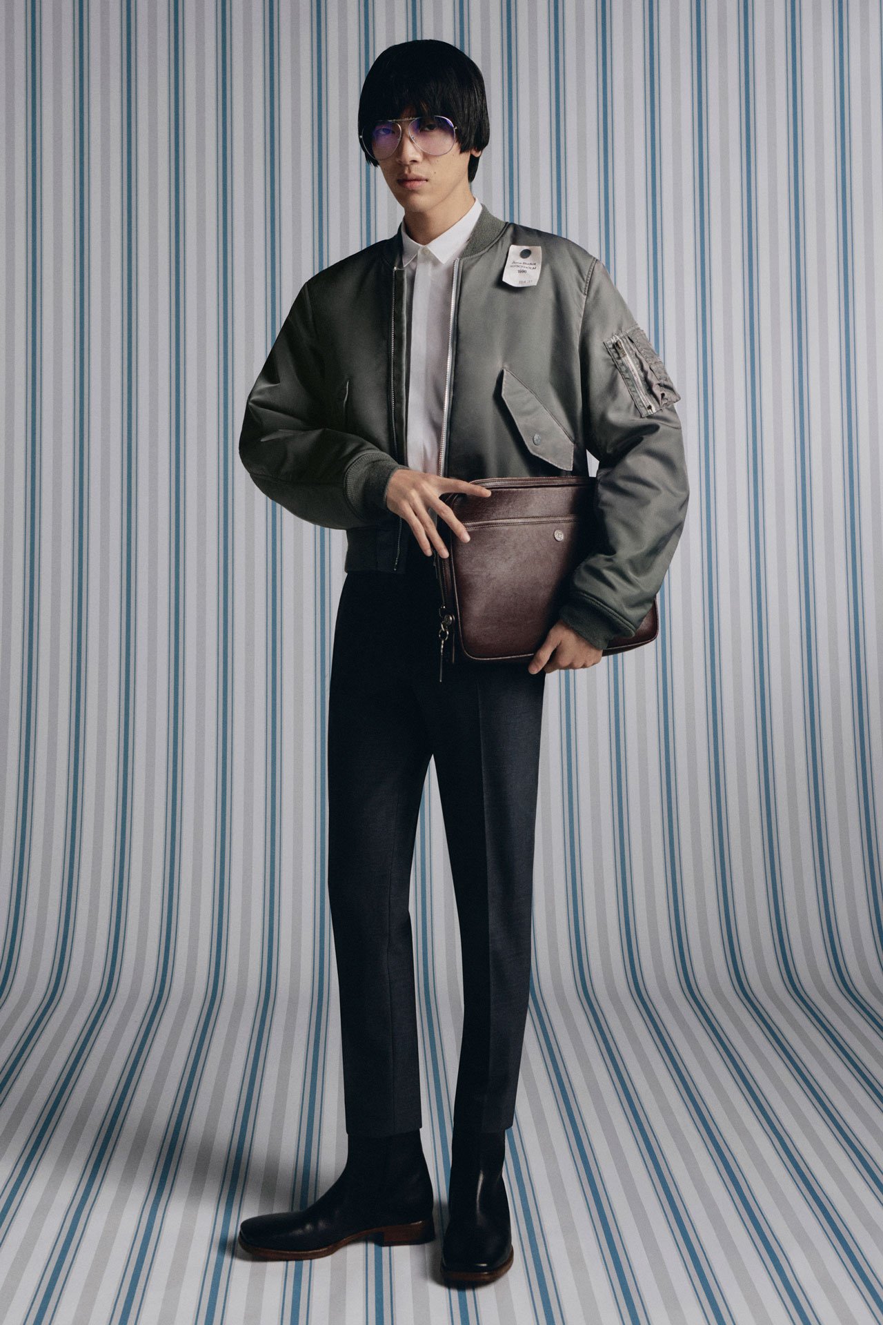 A black haired man in Acne Studios SS26 new arrivals green padded bomber jacket, holding a brown leather laptop case and wearing black tailored pants.