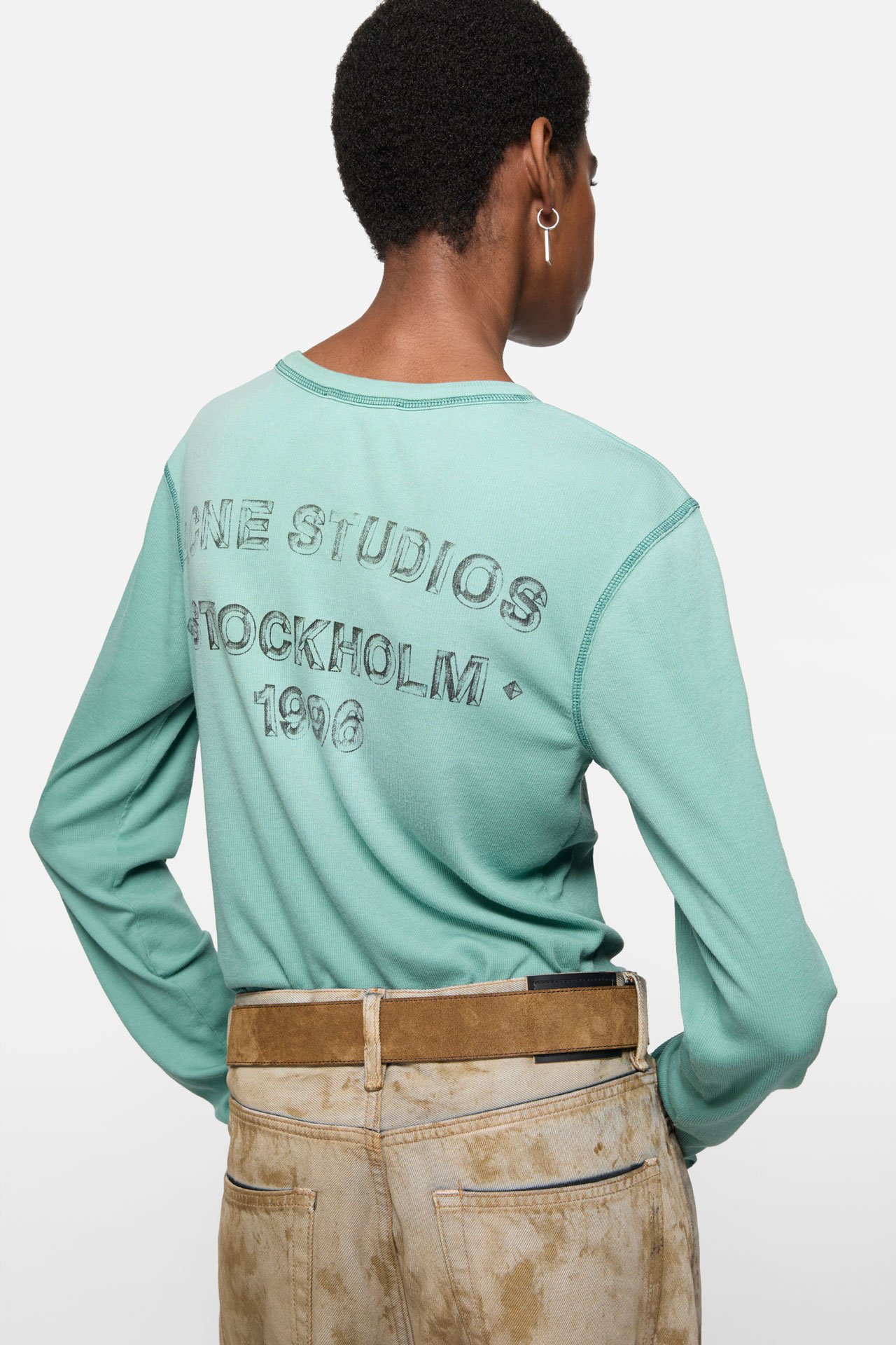 A female model wearing a long-sleeve green t-shirt and beige jeans.