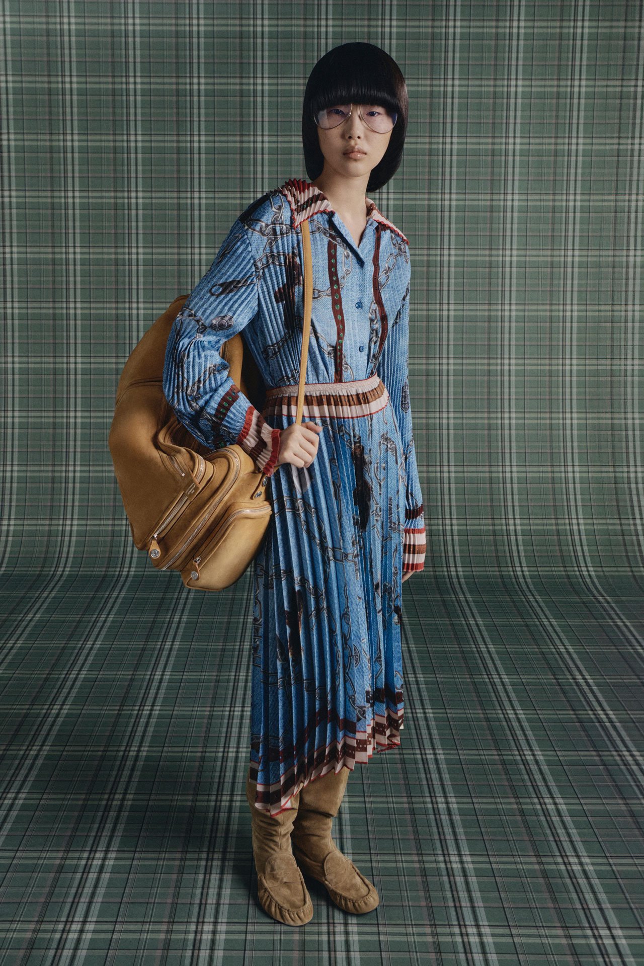 Black short bob-haired female model wearing glasses, Acne Studios blue printed shirt and skirt, beige boots and a big beige Camero bag.
